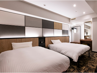 ワシントンホテルプラザのデラックスツインの客室。白いシーツのベッドが2台並び、壁はモダンなデザイン。奥には鏡付きのドレッサーがあり、手前にはソファとテーブルがある。